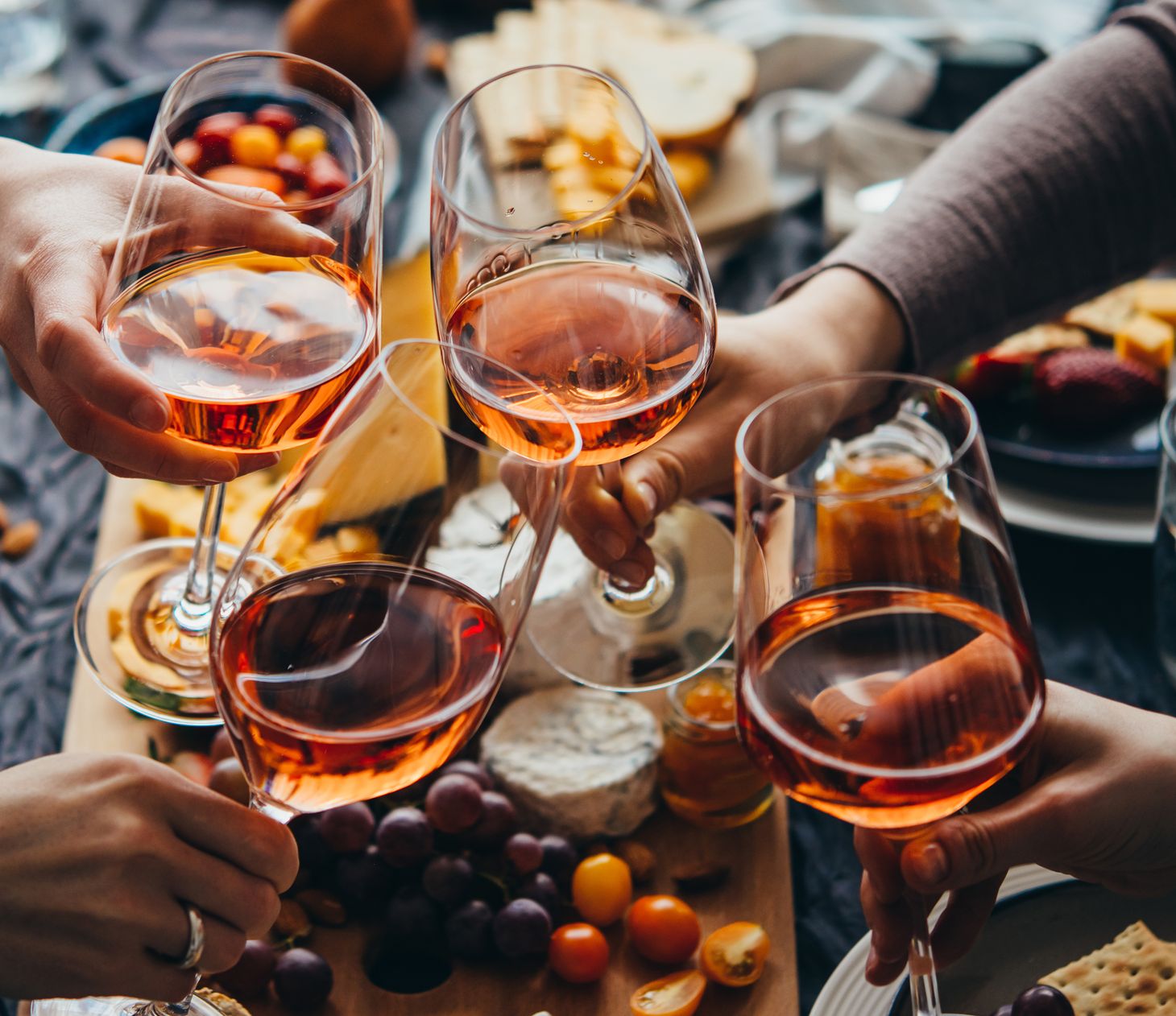 Vier handen proosten met glazen rosé boven een tafel vol kaas en fruit